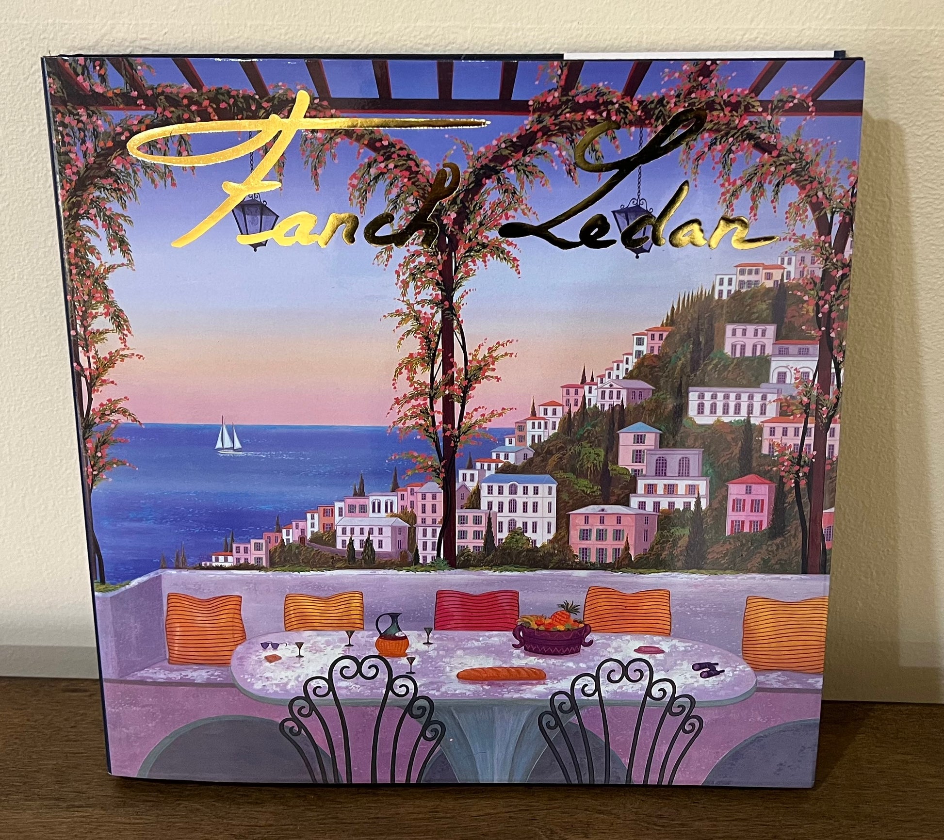 Vibrant-toned book cover with a depiction of a scenic restaurant table overlooking the ocean and pink buildings. The title reads "Fanch Ledan."