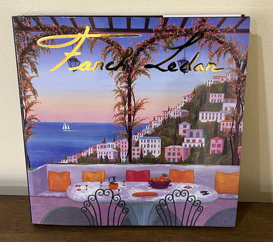 Vibrant-toned book cover with a depiction of a scenic restaurant table overlooking the ocean and pink buildings. The title reads "Fanch Ledan."