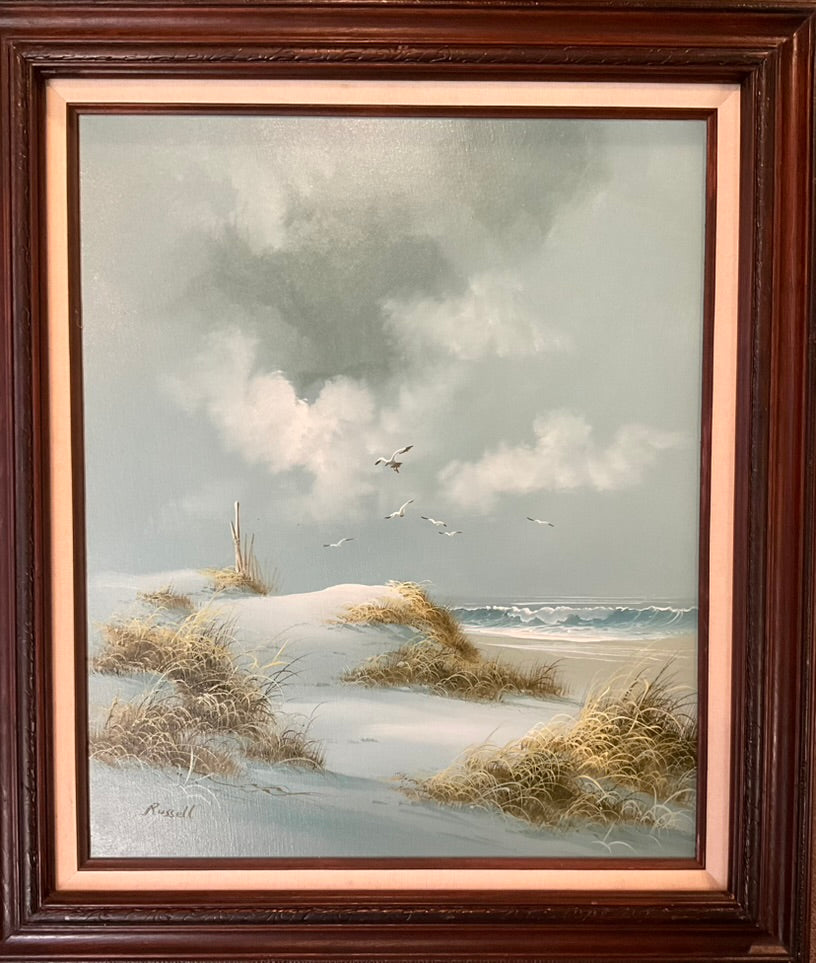 A framed painting of a beach on a cloudy day, featuring, sand grass, seagulls, and an ocean wave crashing to shore.